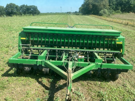 A green John Deere grain drill in the foreground of a field, used for planting cover crops.